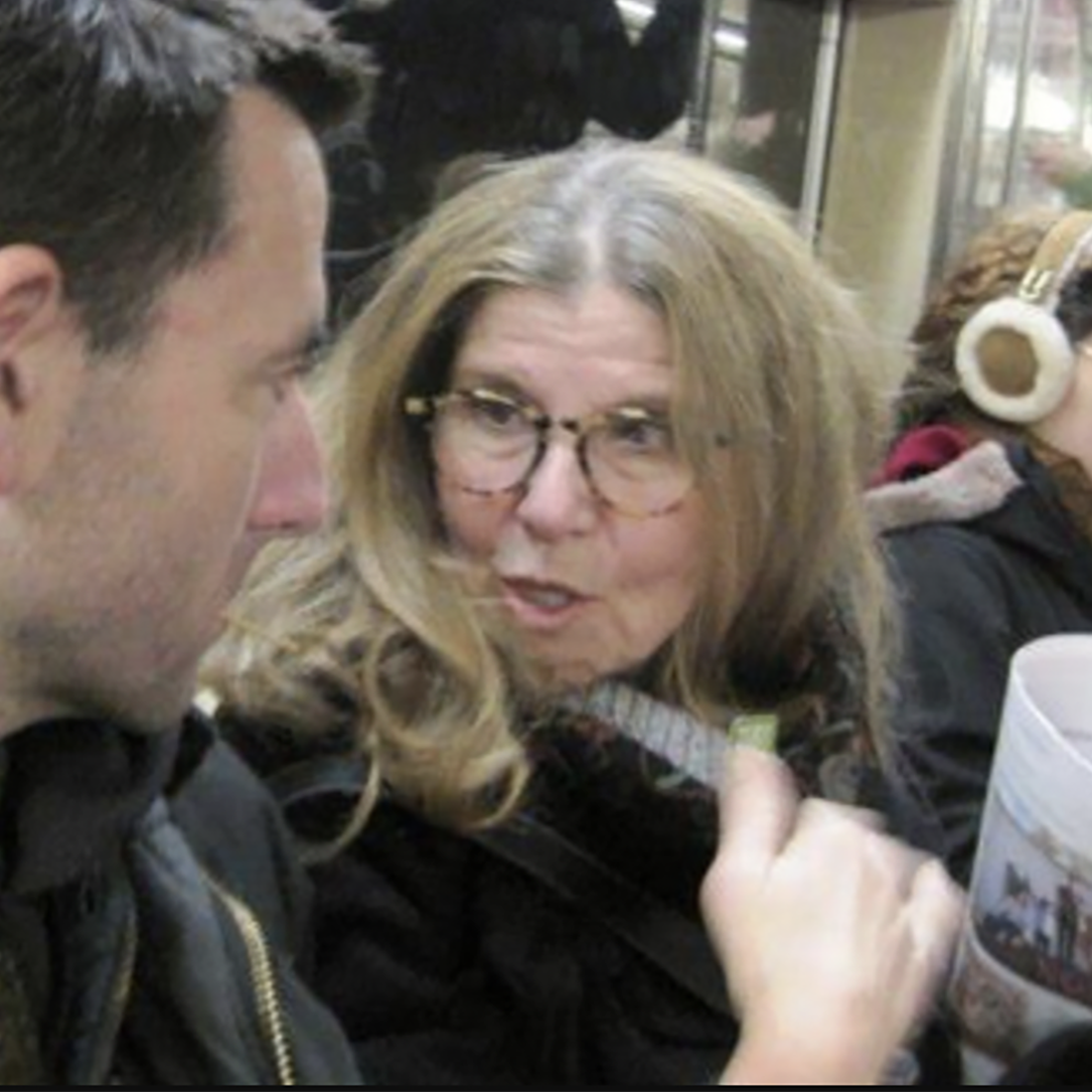 Woman with gray blond hair on street talking to a man with short dark hair.