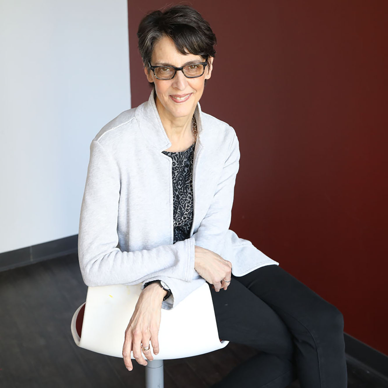 Woman in glasses with dark hair wearing a beige blazer, black top and dark pants, sitting on a stool, legs and are crossed.