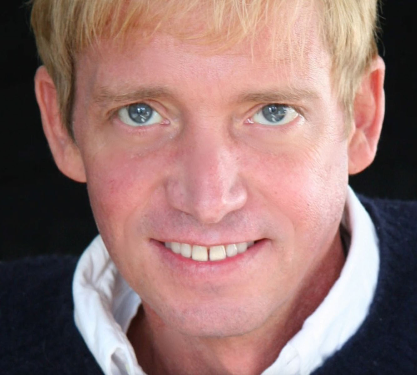 Man with blond hair and blue eyes wering a navy blue sweater and white collared shirt.