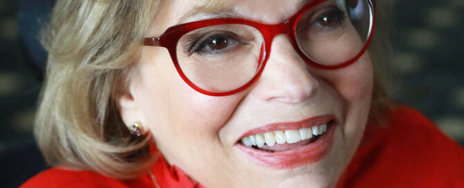 A woman with red glasses, red blouse and light brown hair smiling into the camera.