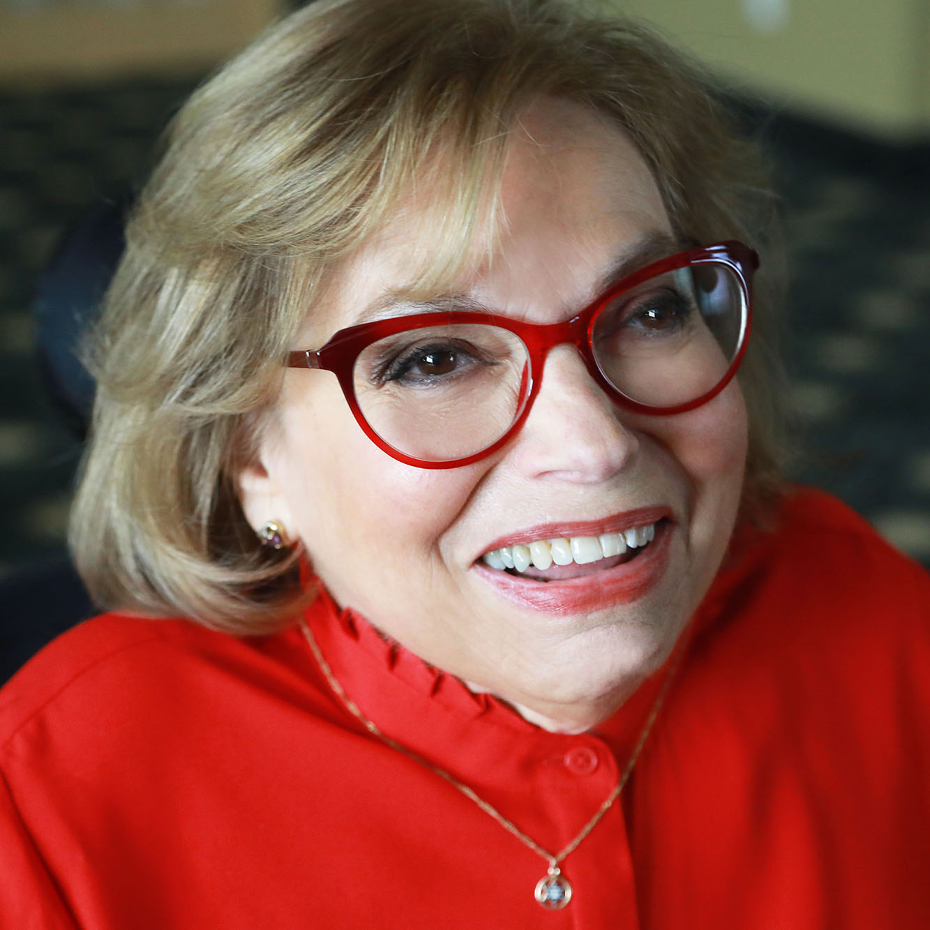 A woman with red glasses, red blouse and light brown hair smiling into the camera.