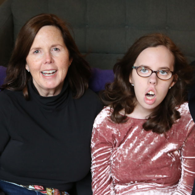 Two women with light skin and long brown hair, wearing black & pink, face the camera.