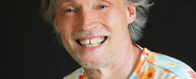 Man with long blong/ gray hair and big smile wearing a floral t-shirt.