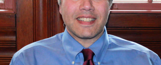 Man with graying hair in blue shirt and dark red tie in front of wood paneling and window.