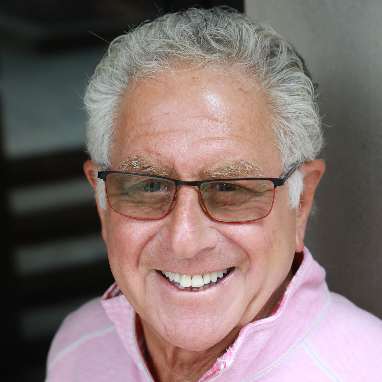 Man with grey hair and glasses, wearing a pink pullover, smiling.