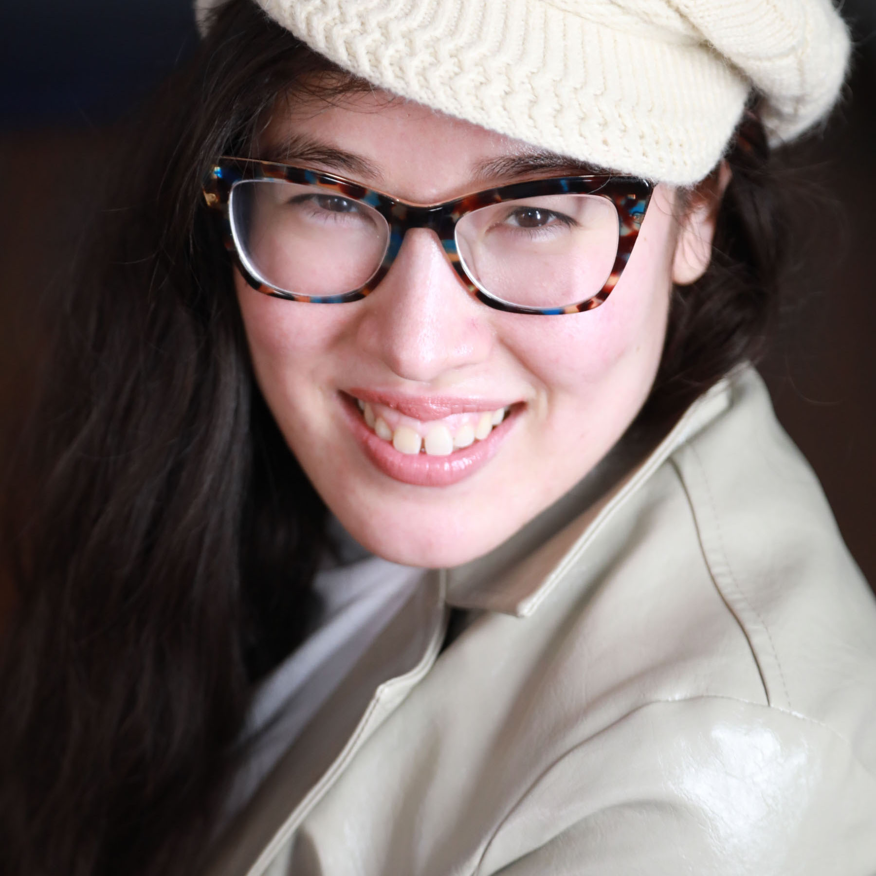 woman in tan leather coat and white wool hat with glasses and dark hair.