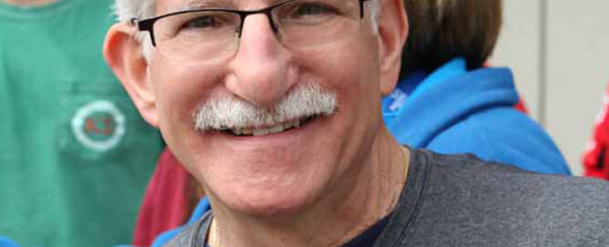Man with white hair and mustache glasses and gray t-shirt.