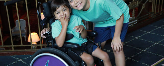 young boy with turquoise shirt in a wheelchair looking into the camera. his brother standing nearby