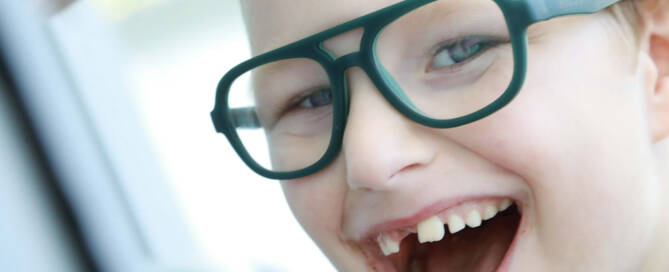 young boy with Angelman Syndrome wearing blue glasses looking into the camera and smiling, closeup photo