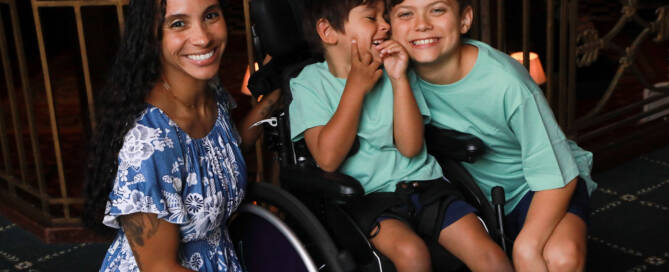 young boy with turquoise shirt in a wheelchair looking into the camera. his brother and mother standing nearby