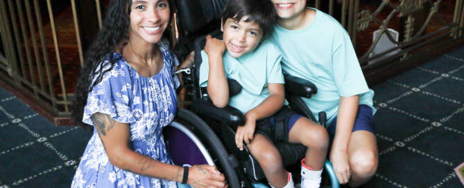 young boy with turquoise shirt in a wheelchair looking into the camera. his brother and mother standing nearby