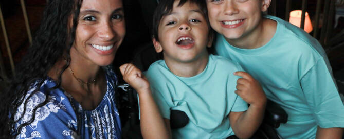 young boy with turquoise shirt in a wheelchair looking into the camera. his brother and mother standing nearby