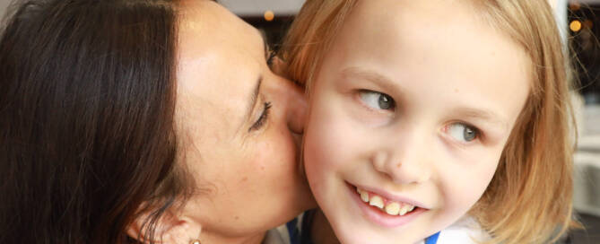 young girl with Phelan-McDermid Syndrome smiling and looking to the right while he mother kisses her cheek