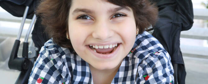 young girl with Angelman Syndrome wearing checkered shirt smiling at the camera