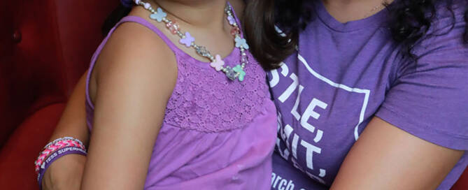 young girl wearing purple shirt being held by her mother wearing purple shirt, both sitting on a red chair