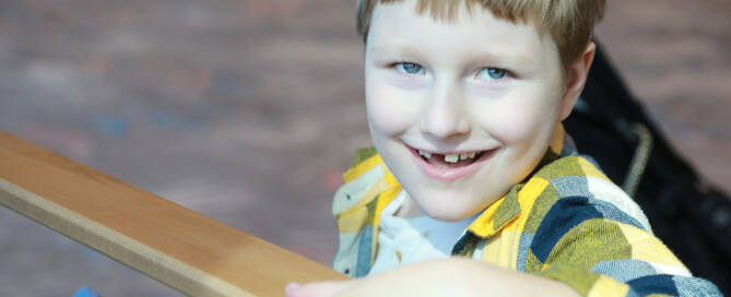 young boy with Angelman Syndrome wearing plaid shirt. Grabbing onto a wooden railing while smiling into the camera