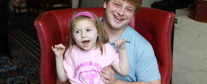 young girl in a pink shirt and pink headband being held by her brother, sitting on a red chair