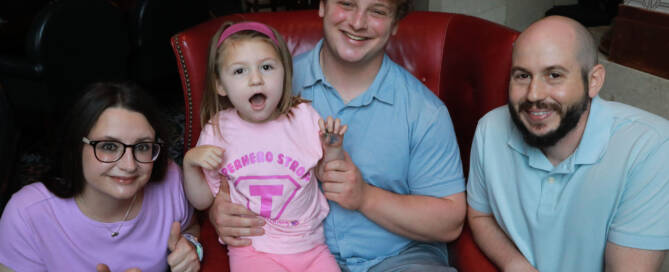 young girl in a pink shirt and pink headband being held by her brother, sitting on a red chair. mother and father crouching on the sides