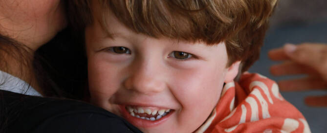 young boy with Phelan-McDermid Syndrome being held by an adult smiles into the camera, closeup photo