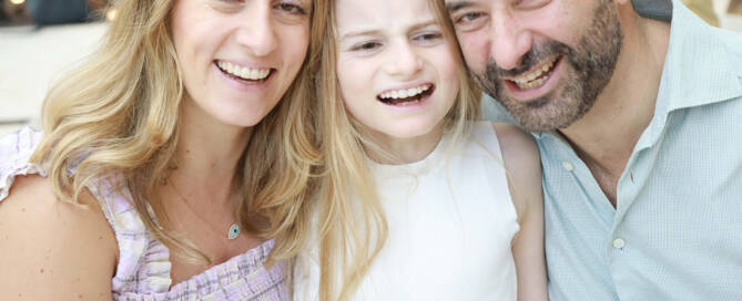 young girl with Angelman Syndrome wearing white shirt, parents on both sides of her. All three smiling