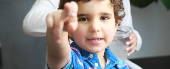 young boy with Phelan-McDermid Syndrome wearing blue button up shirt points at the camera