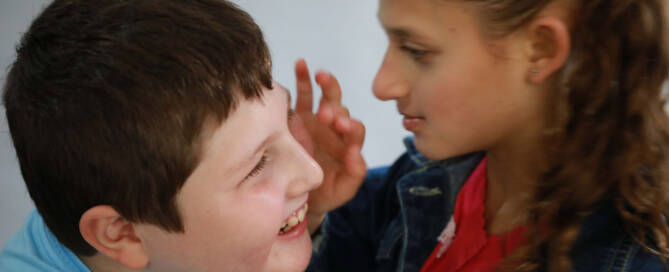 young boy and girl both with Angelman Syndrome. He is smiling while her hand is raised towards his face