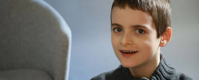 young boy with Angelman Syndrome wearing grey sweater smiling into the camera