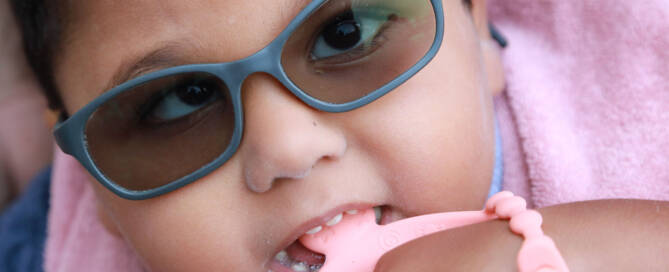 young girl with grey sunglasses and pink outfit looks into the camera while chewing on a pink toy