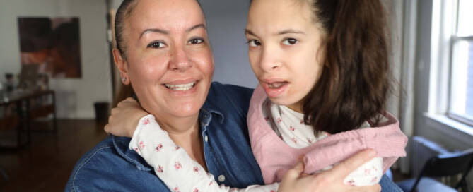 girl with chromosome 5p- (Cri Du Chat) wearing pink and white outfit being held by her mother as they both look into the camera