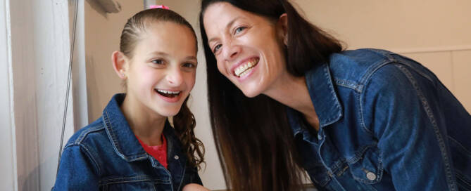 young girl with Angelman Syndrome smiling at the camera while her mom smiles with her