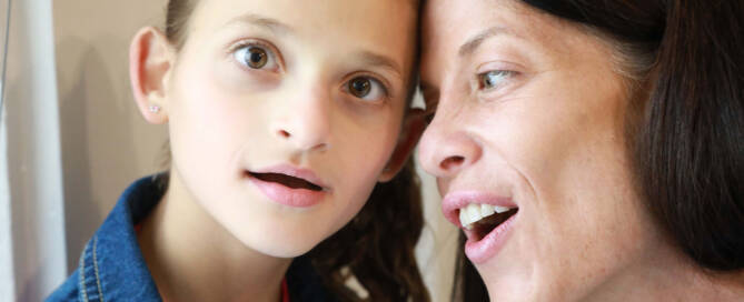 young girl with Angelman Syndrome looking into the camera while her mom is looking at her and smiling