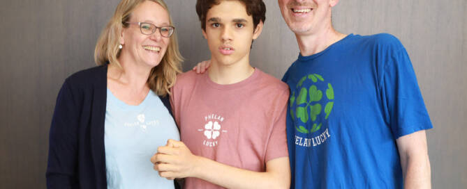 boy with Phelan-McDermid Syndrome wearing light red shirt standing with his mother and father