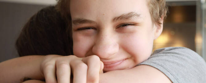 boy with Phelan-McDermid Syndrome wearing grey shirt, smiling while hugging somebody