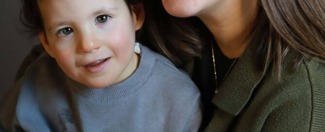 young girl with chromosome 5p- (Cri Du Chat) wearing grey shirt and pink boy sitting in her mother's lap. both looking at camera