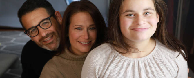 girl with Angelman Syndrome wearing beige shirt smiling into the camera. Her mother and father are behind her, both smiling