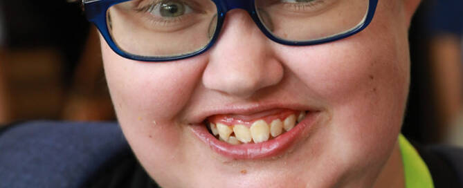 girl with Phelan-McDermid Syndrome wearing navy blue glasses smiles into the camera