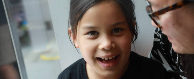 young girl with Phelan-McDermid Syndrome wearing black shirt smiling into the camera, near a window