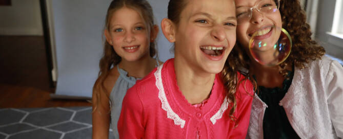 girl with Angelman Syndrome wearing pink shirt laughs at bubbles alongside two other girls