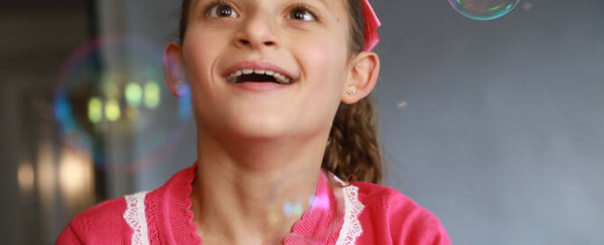 girl with Angelman Syndrome wearing pink shirt looks amazed at bubbles floating around her