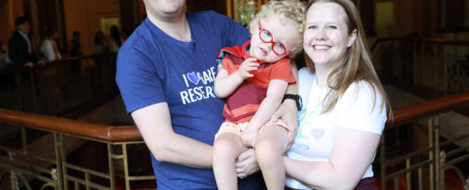 young girl wearing red glasses being held by both her parents, smiling