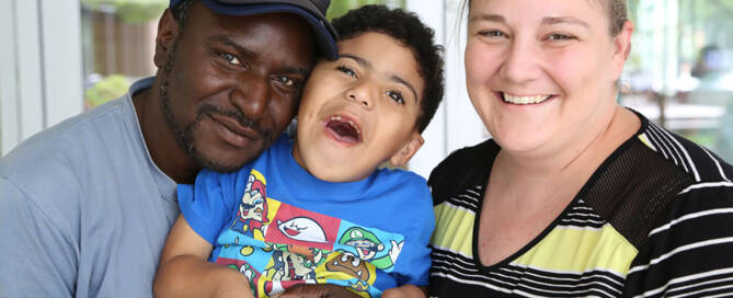 young boy with miller syndrome wearing blue shirt being held by parents and smiling