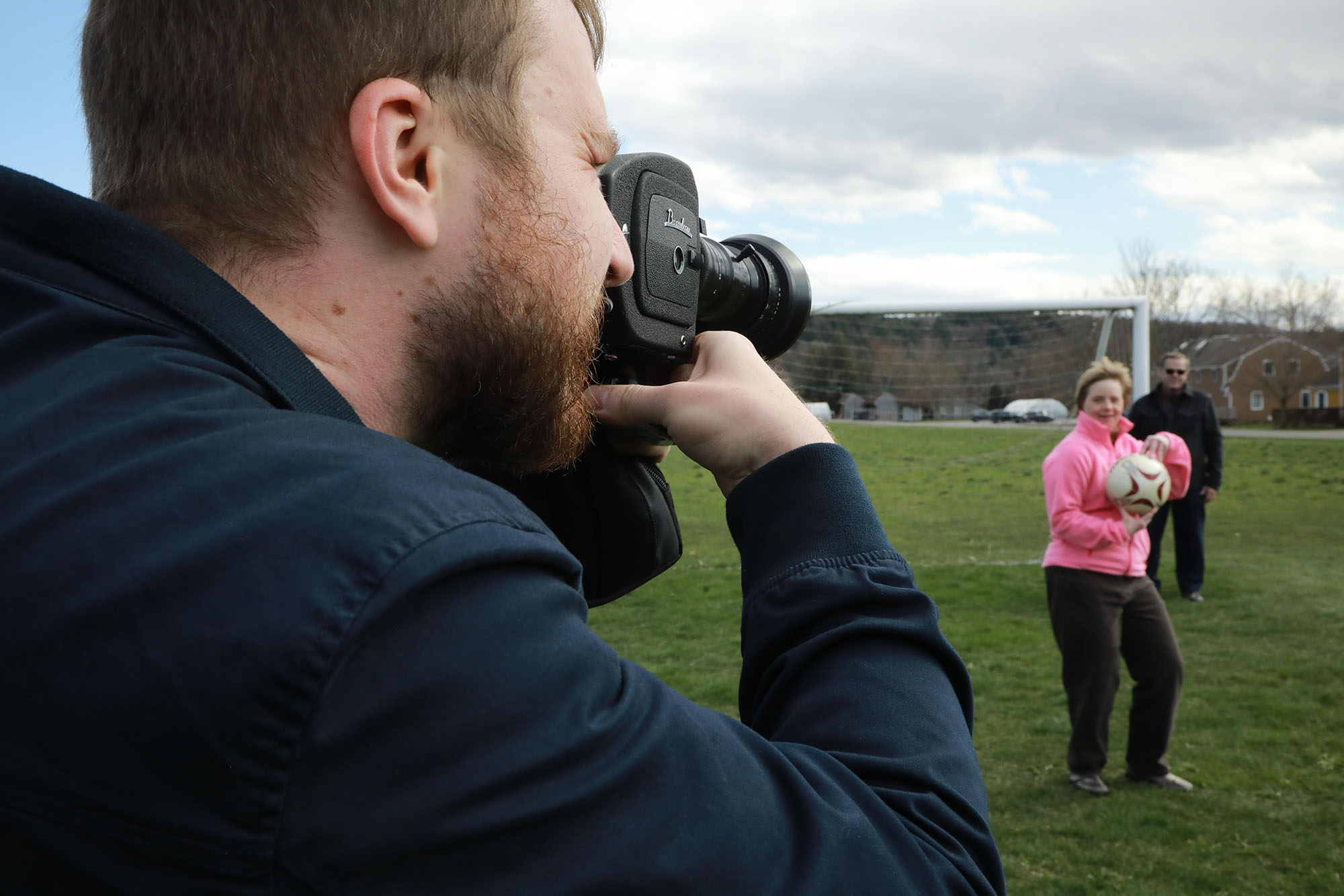 Man holding movie camera and pointing it at woman in pink jacket holding a soccer ball,